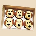A boxed set of six cupcakes decorated with cream and fondant like cute beagle faces with floppy ears and tongues.
