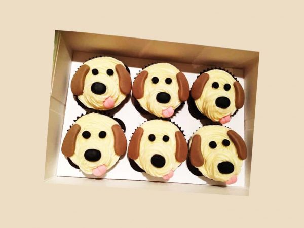 A boxed set of six cupcakes decorated with cream and fondant like cute beagle faces with floppy ears and tongues.