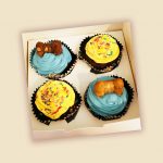 A box of four dog cupcakes with yellow and blue frosting, decorated with colorful sprinkles and bone-shaped treats on top.