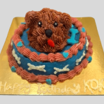 A round two-layer dog cake with a 3D sculpted dog head on top, decorated with pink and blue frosting and small hearts.