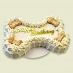 A large bone-shaped dog cake with white frosting, cream piped border, and biscuit decorations on top.