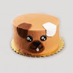 A tall round brown cake featuring a simple cartoon-style dog face made of frosting with cute two ears.