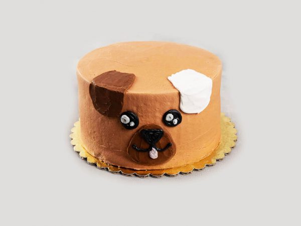 A tall round brown cake featuring a simple cartoon-style dog face made of frosting with cute two ears.