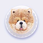 A round cake beautifully decorated to showcase the face of a fluffy Chow Chow dog with textured fur and a pink tongue.