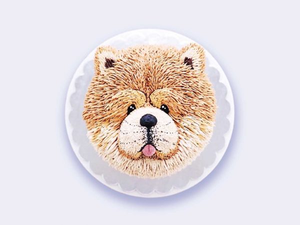 A round cake beautifully decorated to showcase the face of a fluffy Chow Chow dog with textured fur and a pink tongue.