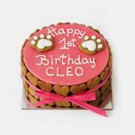 Pink round dog birthday cake with white icing, topped with paw-shaped decorations and heart cookies.