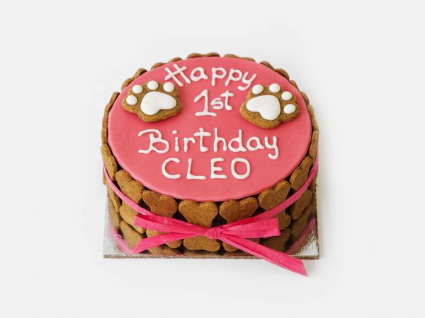 Pink round dog birthday cake with white icing, topped with paw-shaped decorations and heart cookies.