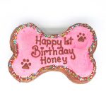 A pink bone-shaped cake covered with colorful sprinkles and decorated with paw shapes on a white base.