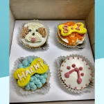 A set of four dog-themed cupcakes in a box, each decorated uniquely with colorful frosting and fondant elements.