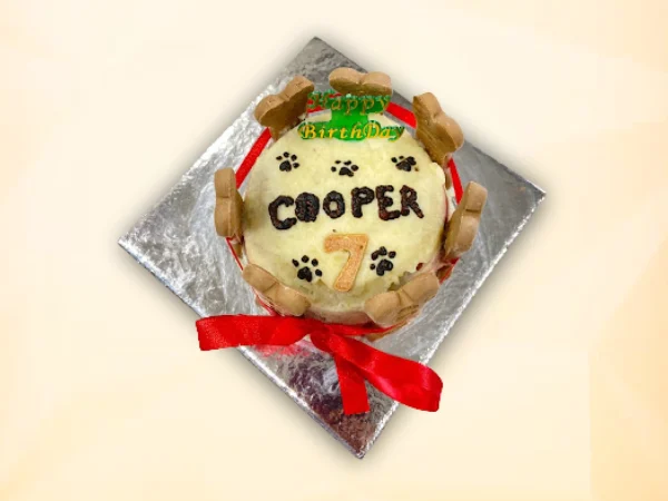 A round dog cake decorated with a red ribbon, bone-shaped biscuits around the sides, and paw prints on top.