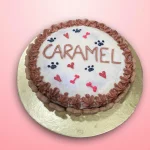 A round white frosted dog cake with a pink cream border, decorated with small paw prints and heart-shaped accents.